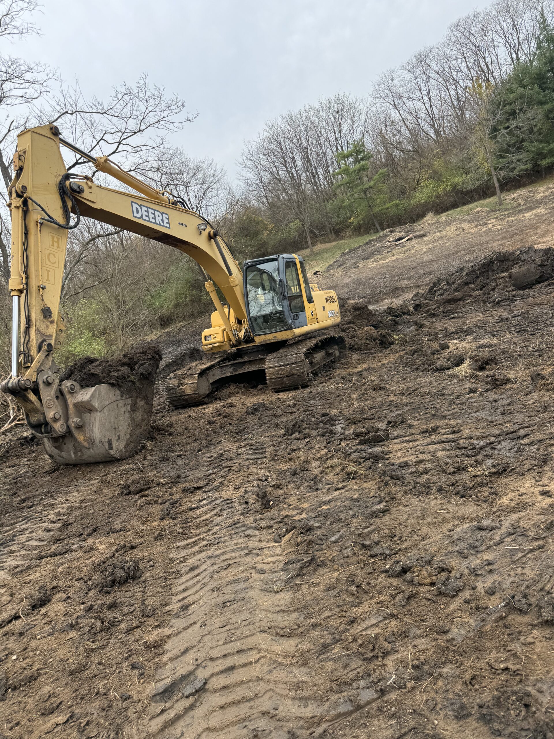A John Deere excavator working on a construction site with disturbed earth and trees in the background.