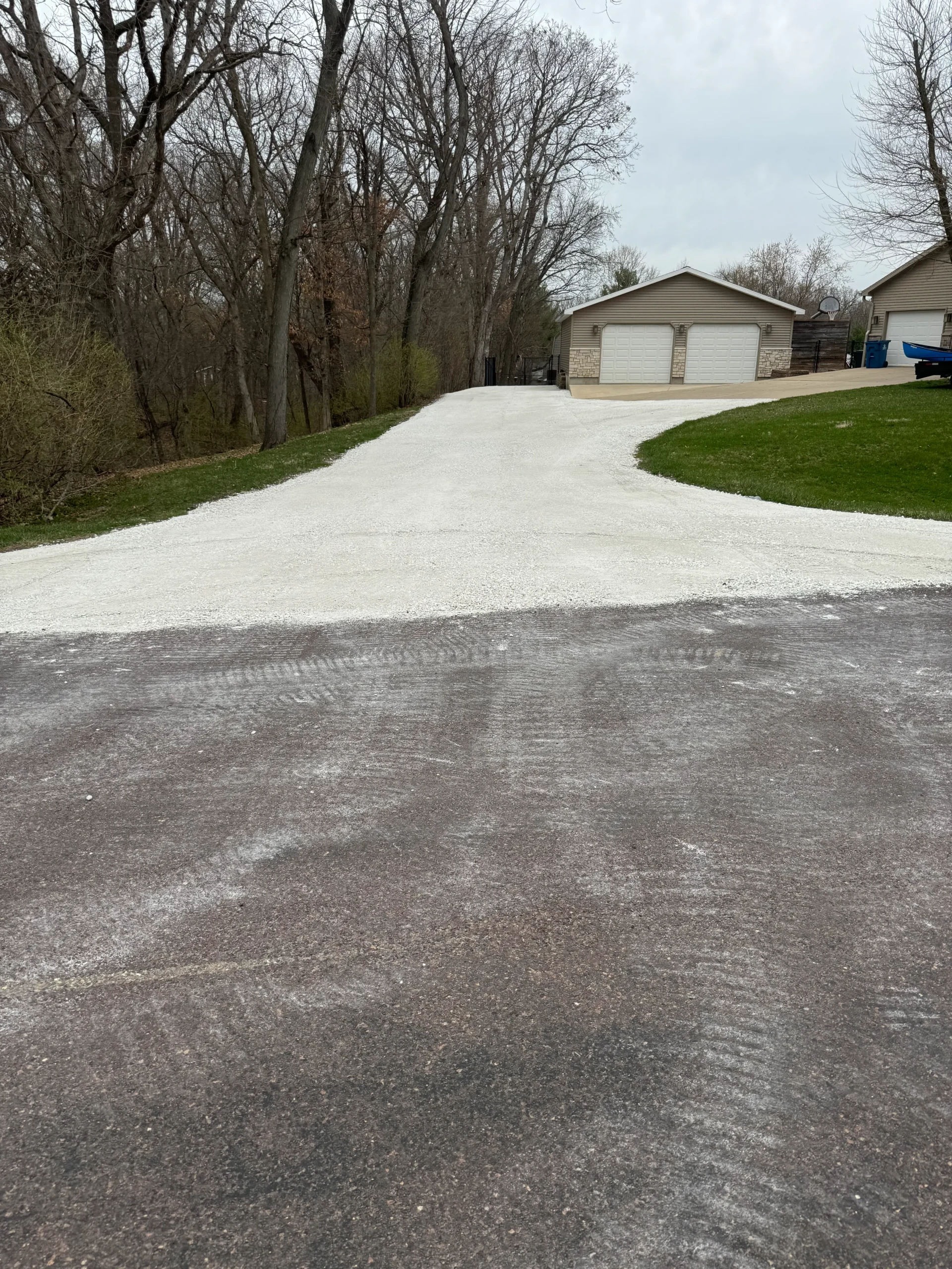 Gravel driveway with a white surface leading to a residential garage in a wooded area.